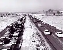 A black-and-white photo shows a four-lane freeway divided by a grass median. In the oncoming lanes, traffic is congested into the distance. With few exceptions, the 401 is surrounded by farmland.