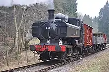 A black pannier tank locomotive is passing along a single track through woodland. The locomotive is pulling a train of a covered red wagon, three grey open wagons, and a red guard's van.