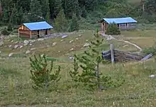 Two log cabins with metal roofs in the distance down a rolling slope. The remains of another are partially visible closer to the camera