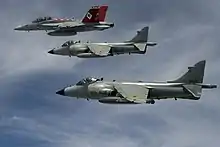 Two similar grey jet aircraft with high-mounted wing flying in formation with another red-tail fighter, which is leading and is furthest from photo. The leading jet is carrying an external fuel tank under fuselage.
