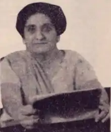 An older South Asian woman wearing a light-colored sari and holding an open book in two hands