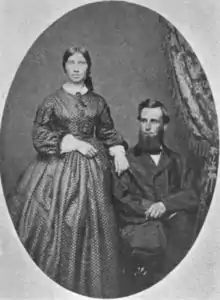 A white couple posed for a portrait in an oval frame. The man is seated, and wears a beard and a suit; the woman is standing, and wears a dress with a high collar and a voluminous skirt. Her hair is center parted; she had one hand on the man's shoulder.