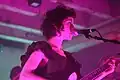 Person with a guitar in their hand by a microphone stand, lit with purple lighting.