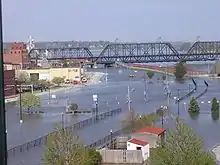 A street with a major amount of water on it due to flooding. A bridge is on the top of the image, and a row of buildings to the left. Sandbags are in front of the buildings