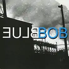 A black-and-white silhouette image of an industrial factory. Large volumes of smoke are visible in the sky. Uppercase white mirrored text in the center reads "Blue" (stylized as "ƎU⅃ᗺ"); uppercase blue text next to it reads "Bob", with black outline around the text.