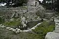 The pool of the sacred spring with the stepped retaining wall (L), the temple podium and the church of Hagios Georgios at center