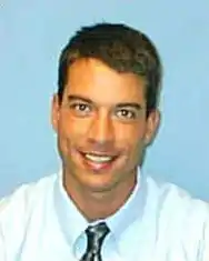 A smiling clean-shaven young man with short dark hair wearing a white shirt and silvery tie against a blue background