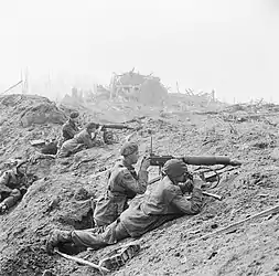Five men with two guns in a devastated landscape. The remains of a bombed town are in the background