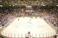 The view of an ice hockey rink from behind and above one goal. Most players, dressed in either red and white or blue and black, are in the far right corner of the rink. Above center ice an electronic scoreboard reads "9:09" in green; under that, red numbers read from left to right "0", "1", and "0". The "1" is smaller than the two "0"s. On the visible end of the rink, a red sign reads in black and white text "Pizza Hut".