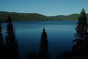 A large lake under blue skies surrounded by forested hills with steep slopes under a clear blue sky