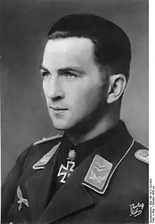 The head of a young man, shown in semi-profile. He wears a military uniform with an eagle above his right breast pocket, an iron cross is displayed at the front of his shirt collar. His hair is dark, short and combed to the back, his nose is long and straight, and his facial expression is determined; looking to the right of the camera.