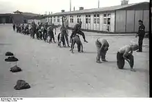 A line of half-naked prisoners performing "leap frog", under supervision of one of the Kapos. In the background the main gate to Mauthausen as well as two wooden barracks are visible.