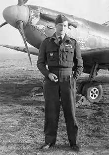 A full length picture of a man in RAF uniform. He stands in front of a parked aircraft, with one hand in a pocket and holding a pipe in the other.