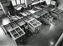 Looking down from above at a room at an intricate mechanical device which fills the room. In the background, a man sits at a desk next to a filing cabinet. Owens 1991, p.&nbsp;15.