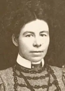 A young white woman with dark hair center parted and dressed up from the shoulders; wearing a high-collared dress with braid trim and puff sleeves