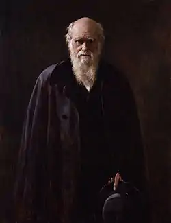Three-quarter portrait of a senior Darwin dressed in black before a black background; his face and six-inch white beard are dramatically lit from the side;  his eyes are shaded by his brows and look directly and thoughtfully at the viewer