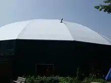 A round, green building with a white tent-like roof stands against a clear summer sky.