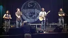 A woman and three men on stage; the woman with a ukulele and one of the men with a guitar