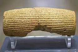 Rear view of a barrel-shaped clay cylinder resting on a stand. The cylinder is round with square edge very close up. It is covered with lines of cuneiform and has a hollow centre. The centre is lined with plate measuring around 10 millimeters in thickness.text
