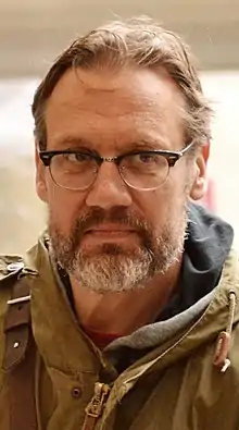 Middle-aged white male with short hair and facial hair, wearing brown jacket and glasses, looking left of camera