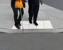 Two people stand on top of light-colored truncated domes at a curb cut.  They both face the parking lot, and the person in front holds a white cane.