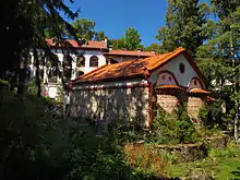Apse view of a late medieval stone church with a tiled roof and a residential building in the background, all located in a forested area