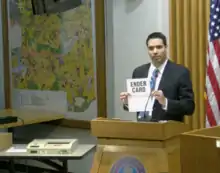 Man holds up a paper labeled "ender card" next to a machine that processes ballots and tabulates results.