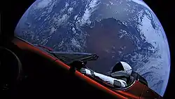Large circular disc of a fully-illuminated planet Earth showing Australia floating in the blackness of space. In front of Earth is a red convertible sports-car seen from the side. A humanoid figure wearing a white-and-black spacesuit is seated in the driving seat with the right-arm holding the steering wheel, and the left-arm resting on the top of the car door.