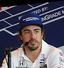 A man in his mid-30s wearing a navy blue baseball cap with a team name in white capital letters and a white T-shirt with sponsors logos that is unbuttoned at the top button . He is sporting a stubble.