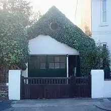 An extremely small workshop adjoining a house and standing behind a wooden gate with white gateposts. It has green double doors with an open door to their right.  Above this is a white-painted wall which is heavily draped with ivy, although a circular louvre is clearly visible through this.  The roof is steep and pointed.