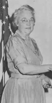 An older white woman with white hair, wearing glasses and a light-colored dress in front of an American flag