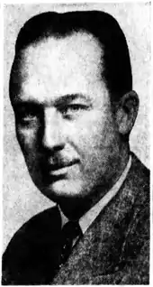 Black-and-white photographic portrait of a smiling adult man with slicked-back hair, a toothbrush mustache, and well-defined facial features. He eyes the camera with a classically handsome confidence. He is depicted wearing a suit.