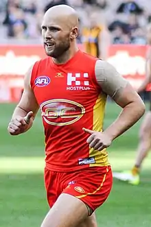 A male athlete with bald head wearing a sleeveless jersey and shorts jogs on the grass surface of the playing arena.