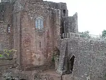 A castle, with a flat fronted tower facing the viewer with a stained glass window in the middle of it; a stone causeway is on the right of the picture, leading to a gateway to the right of the tower – a partially filled arch is supporting the causeway.