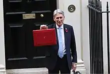 Colour photograph of Philip Hammond holding up his red ministerial box outside 11 Downing Street in October 2018