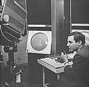 A young man sits holding a microphone in his left hand while manipulating the console of an apparatus with his right. To his left a large television camera is trained on a large, circular cathode ray tube display.
