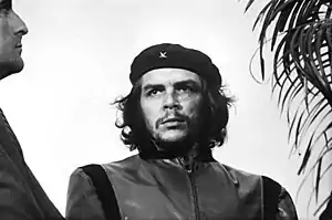 Black-and-white image of a man looking to his right. He wears a black beret with a little "x" and a closed jacket. His hair is shoulder-length, curly and black. On his right there is a man looking to him and on his left, there is a plant.