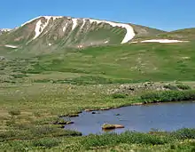 A grassy yet barren landscape with a small pond in the right foreground and distant ridgeline and mountains in the background