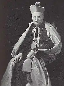 A bespectacled man holding a crosier in his left hand and wearing a mitre and liturgical vestments faces forward.