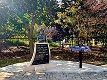 A monument made of bronze and limestone, with a descriptive table to the right with a background of fall foliage.
