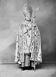 A bespectacled man holding a crosier in his left hand and wearing a mitre and liturgical vestments faces forward.