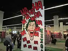 A large playing card bearing the Joker's face stands before a series of art works featuring the Joker