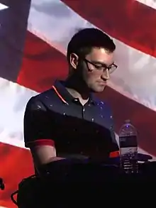 A white man with glasses; near him is an audio equipment, and on the background is the American flag