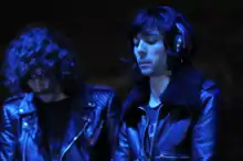 Two men, dressed in black, under a blue light