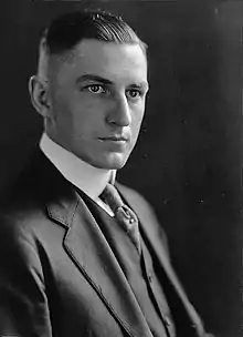 A man in his late twenties with dark, short, slicked-back hair and wearing a black coat and tie and white shirt