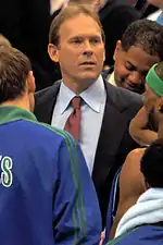 A man, wearing a black suit, blue shirt and red tie, is standing in the middle of a huddle.