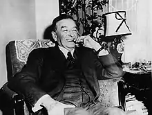 A middle-aged man in a suit sitting in a chair and smoking a cigar