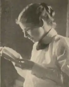 A young white woman, hair in a bun, wearing a high-collared white blouse, holding and regarding a small book or pamphlet