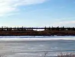 A frozen river passes through flat country. Short trees grow on the riverbanks; tall mountains are in the far distance.