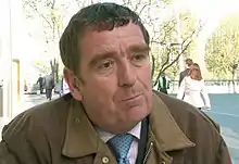 A man in a brown jacket, shirt and tie is pictured sitting outdoors in late spring